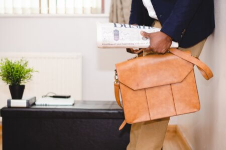 man holding brown leather bag leaning on white wall