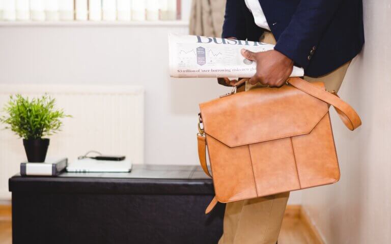 man holding brown leather bag leaning on white wall